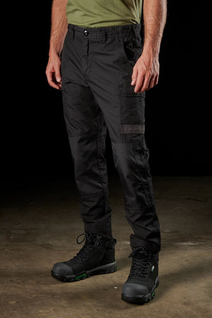 Full-body view of a person wearing the FXD WP-5 black cargo pants made from advanced stretch fabric and black work boots with green accents, standing against a plain dark background. The person is also wearing a green shirt, though only the lower part is visible. The setting appears to be indoors.