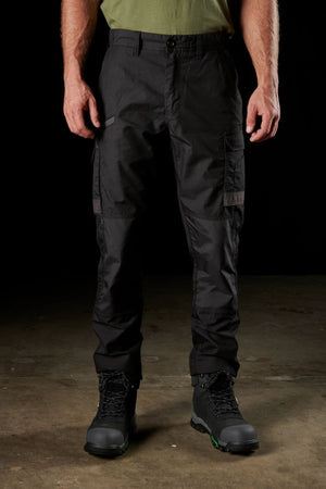 A person standing on a concrete floor, wearing a green shirt and black FXD WP-5 cargo pants crafted from advanced stretch fabric. The black work boots featuring green accents complete their look, set off by the dark background that emphasizes the lower part of their body.