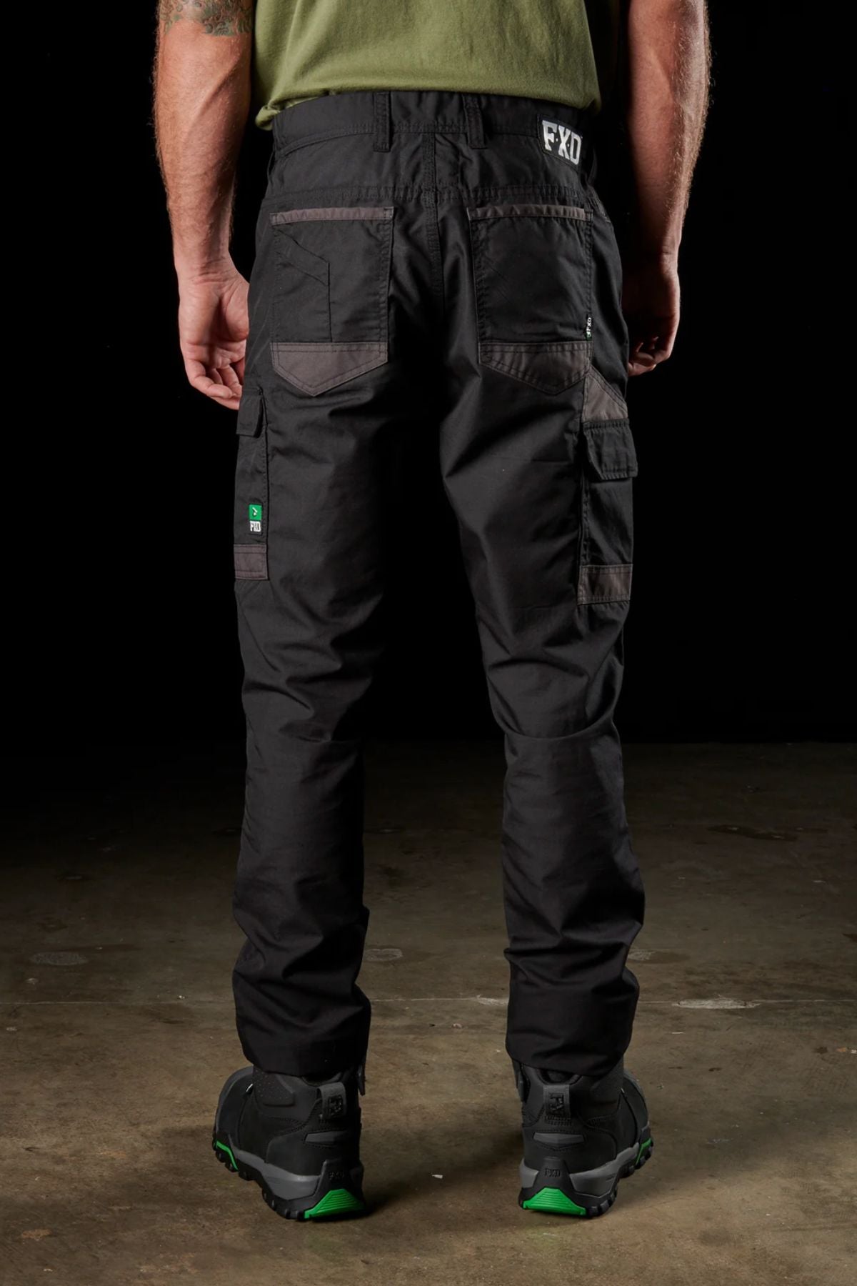 A person stands with their back to the camera, wearing black FXD WP-5 work pants made from advanced stretch fabric and equipped with modern utility pockets. The pants display a small green logo on the back left pocket and "FXD" lettering on the right side. The person also wears dark shoes adorned with green accents.