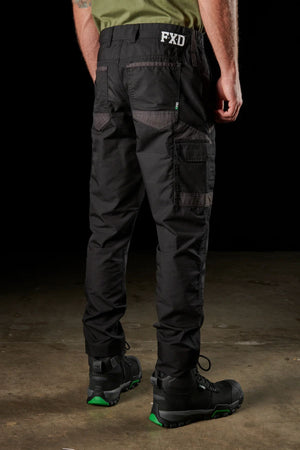 A person wearing black FXD WP-5 work pants, featuring the "FXD" text on the back pocket, is standing on a concrete floor. They have paired the pants with black work boots accented in green and are also sporting a green shirt tucked into the pants. The FXD WP-5 work pants boast advanced stretch fabric and modern utility pockets, enhancing both comfort and functionality. The image captures this scene from behind.