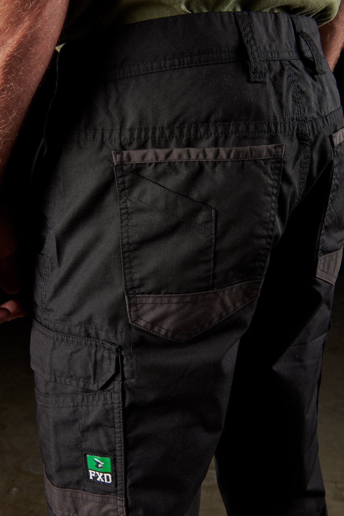 Close-up view of a person's lower body wearing FXD WP-5 black work pants, crafted from advanced stretch fabric. These pants feature modern utility pockets, intricate stitching details, and a small green FXD WP-5 logo patch near the left knee. The person is paired with a green shirt.