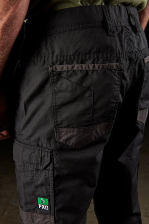 Close-up view of a person's lower body wearing FXD WP-5 black work pants, crafted from advanced stretch fabric. These pants feature modern utility pockets, intricate stitching details, and a small green FXD WP-5 logo patch near the left knee. The person is paired with a green shirt.