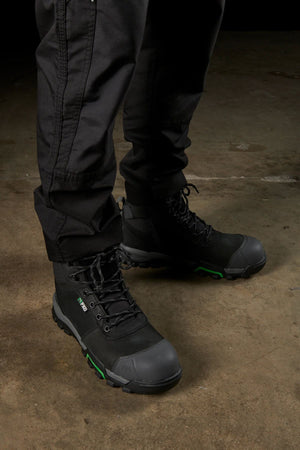 A person wearing black tactical pants featuring modern utility pockets stands on a concrete surface, complemented by the FXD WP-5 rugged work boots, which are lace-up with reinforced toes and thick soles designed for heavy-duty use. The boots feature distinct green accents.