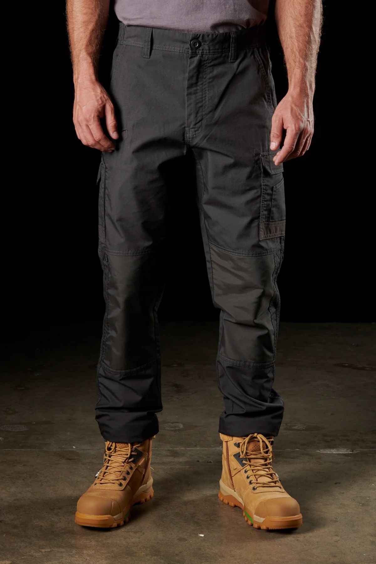 A person wearing a gray shirt and FXD WP-5 dark tactical cargo pants stands on a concrete surface, with tan lace-up combat boots. The ultra-durable FXD WP-5 clothing, complemented by advanced stretch fabric, is highlighted against the black background.