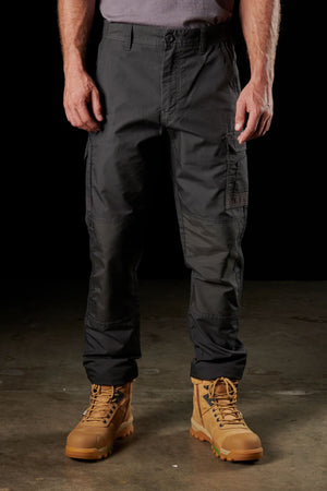 A person wearing a gray shirt and FXD WP-5 dark tactical cargo pants stands on a concrete surface, with tan lace-up combat boots. The ultra-durable FXD WP-5 clothing, complemented by advanced stretch fabric, is highlighted against the black background.