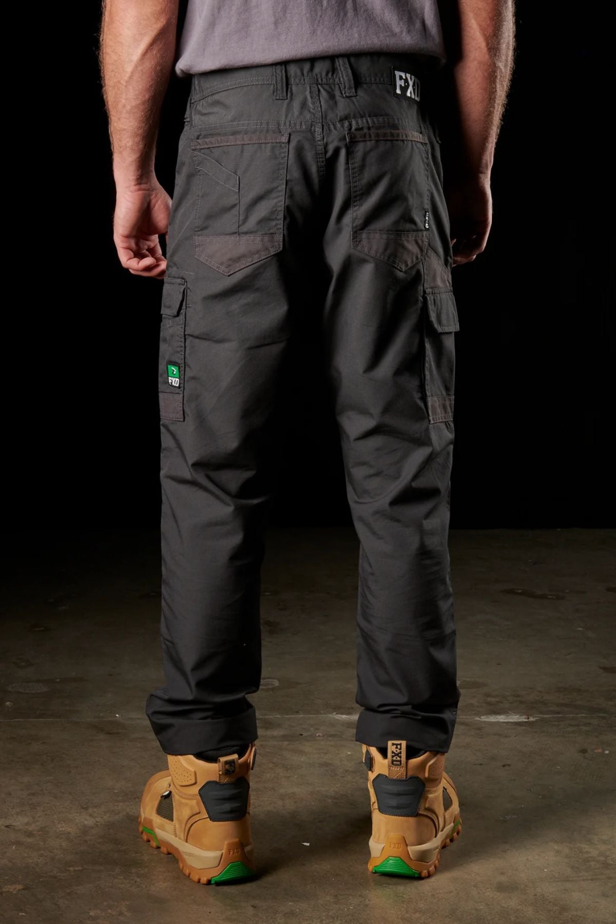 The scene features a man viewed from behind, dressed in FXD WP-5 dark cargo pants made of ultra-durable, advanced stretch fabric and a gray shirt. His legs are straight as he stands on a concrete surface, wearing rugged tan work boots with green soles against a dark background.