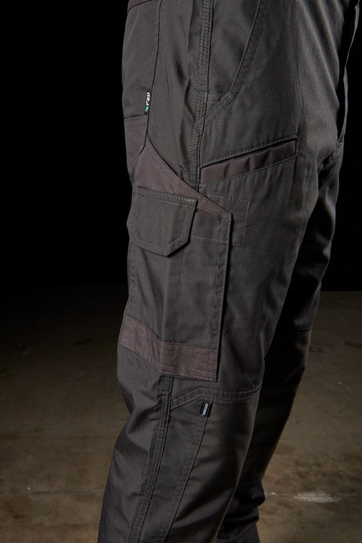 Side view of a person wearing FXD WP-5 cargo pants in a dark gray or black color, with emphasis on the pockets and stitching details. The background is dark, highlighting the ultra-durable texture and design of the pants. Made with advanced stretch fabric for ultimate breathability, the person stands on a concrete floor.