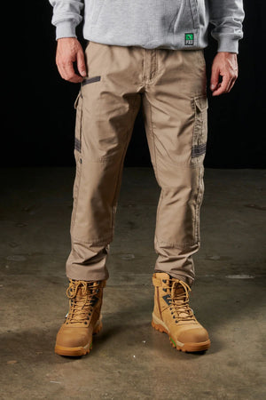 A person wearing beige FXD WP-5 cargo pants, brown work boots, and a gray hoodie stands on a concrete floor. The pants feature modern utility pockets and zipper details. The image is cropped at the waist and ankles, focusing on the lower half of the body.