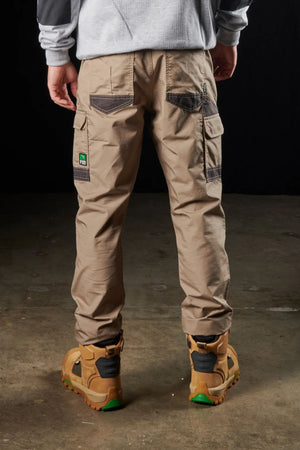 A person wearing khaki utility pants featuring modern utility pockets and advanced stretch fabric, along with a green and white patch on the left pocket, stands on a concrete floor with their back to the camera. The individual is also sporting light brown FXD WP-5 work boots accented in green and orange.