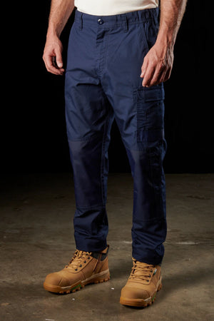 A person stands against a dark background, wearing blue FXD WP-5 work pants with multiple pockets and tan work boots. The FXD WP-5 pants, crafted from advanced stretch fabric for ultimate breathability, feature reinforced knee patches. The sturdy-soled boots are ideal for outdoor or industrial work.