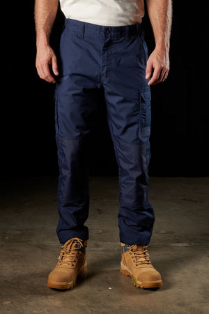 A person stands against a dark background, wearing dark blue FXD WP-5 cargo pants made with advanced stretch fabric and light brown work boots. With their arms at their sides, a white shirt is partially visible at the top of the image.