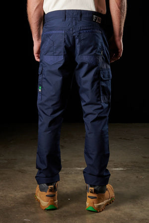 A person wearing dark blue FXD WP-5 cargo pants, made from advanced stretch fabric, stands facing backward, showcasing the back pockets and side utility pockets. They are also wearing tan and green work boots with visible tread patterns on a concrete floor, set against a dark background.
