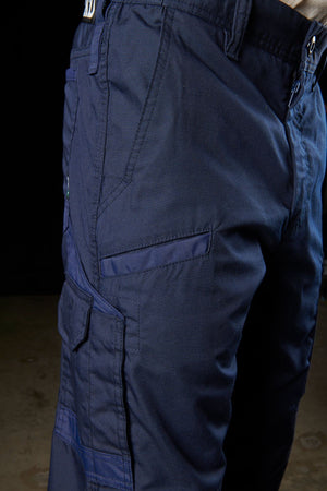 A close-up of a person wearing FXD WP-5 cargo pants in navy blue showcases the multiple pockets on the left side, including a flap pocket and a zippered pocket. The durable advanced stretch fabric is clearly visible, set against a dark background that makes the pants stand out prominently.