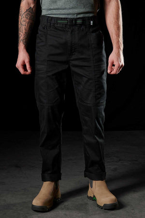 A person in FXD WP-6 black work pants with a stretch waistband, a grey t-shirt, and tan utility boots stands against a dark backdrop, revealing tattoos on one arm.