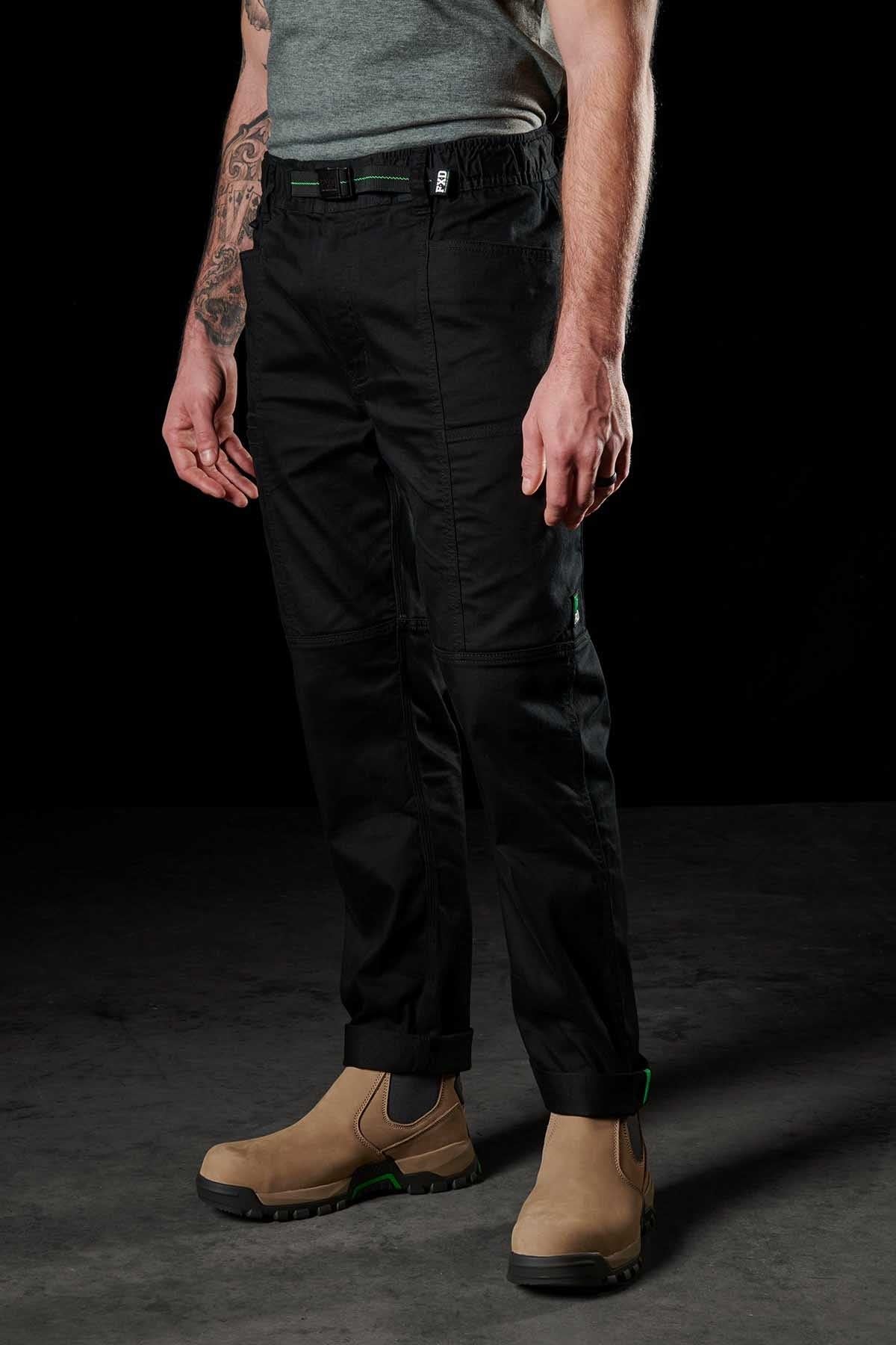 A person in FXD WP-6 black work pants, featuring a stretch waistband for comfort and flexibility, stands against a dark background. The pants are slightly rolled up at the ankles. They're also wearing tan boots and a gray t-shirt, with a visible arm tattoo.