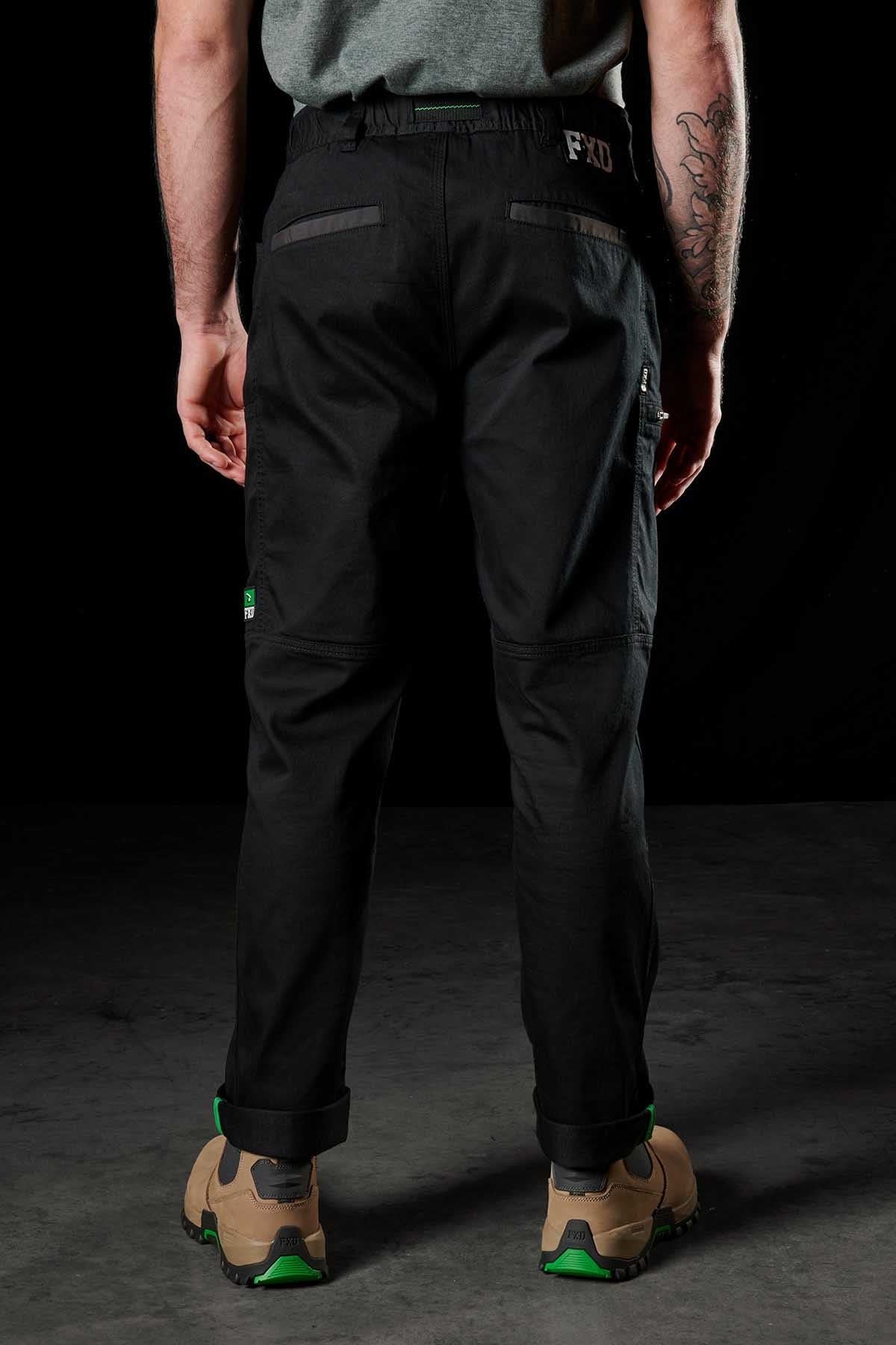 A person with a visible arm tattoo wears FXD WP-6 black work pants featuring a stretch waistband and reinforced patch pockets, paired with a green belt and tan hiking shoes with green accents, standing against a dark background.