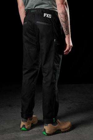 Against a dark background, a person with a tattooed arm stands wearing black FXD WP-6 work pants featuring a stretch waistband and tan work boots.