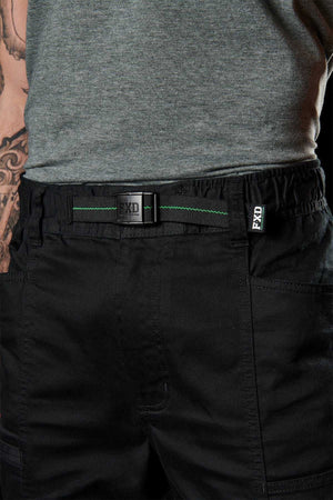 A close-up shows a person wearing FXD WP-6 work pants with a stretch waistband and green-threaded belt. They are paired with a grey t-shirt, and part of the person's left tattooed arm is visible.