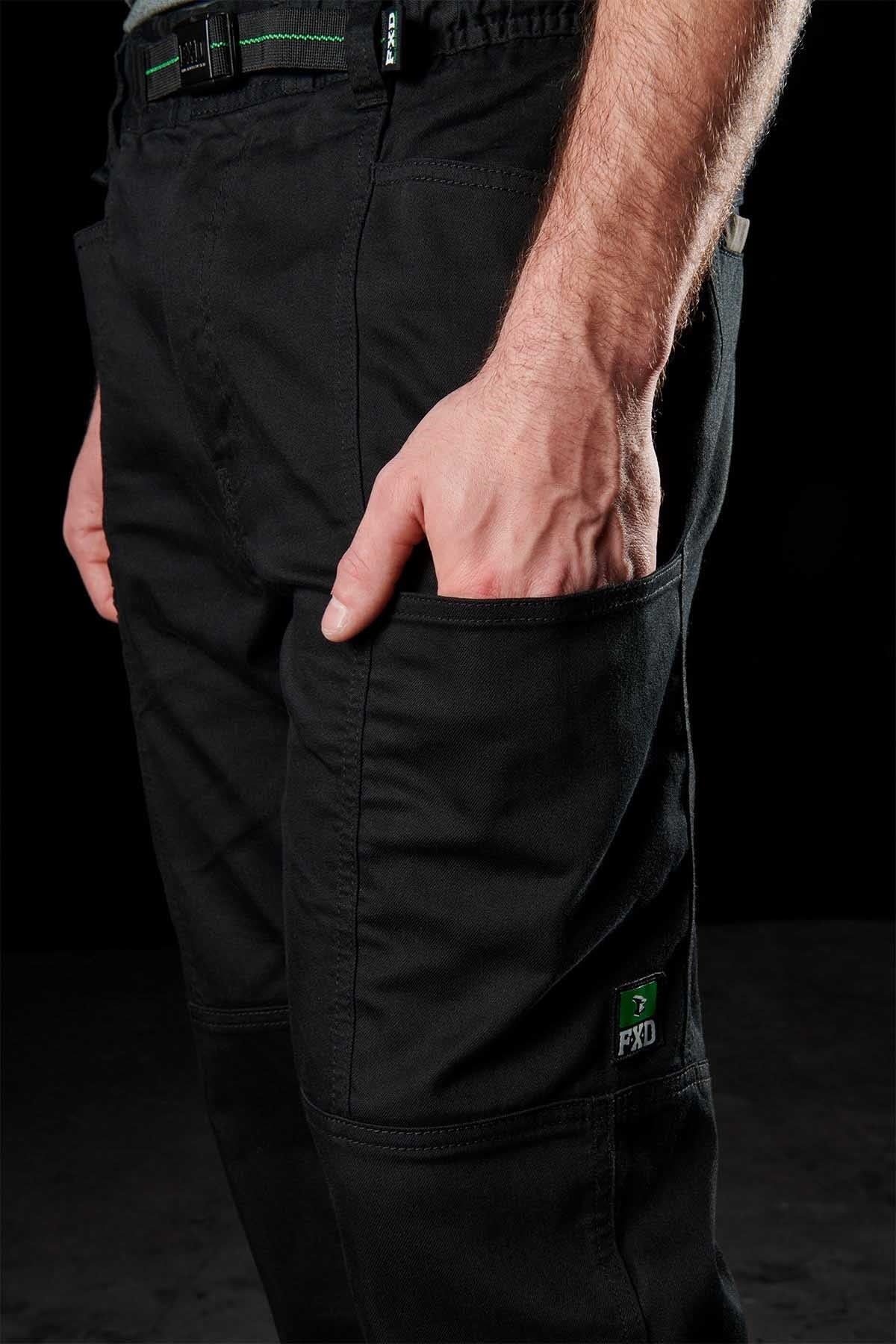 A person models FXD WP-6 black work pants with a stretch waistband and reinforced knee patches, their right hand in a pocket. A small green logo on the leg stands out against the dark background, highlighting the pants' features.