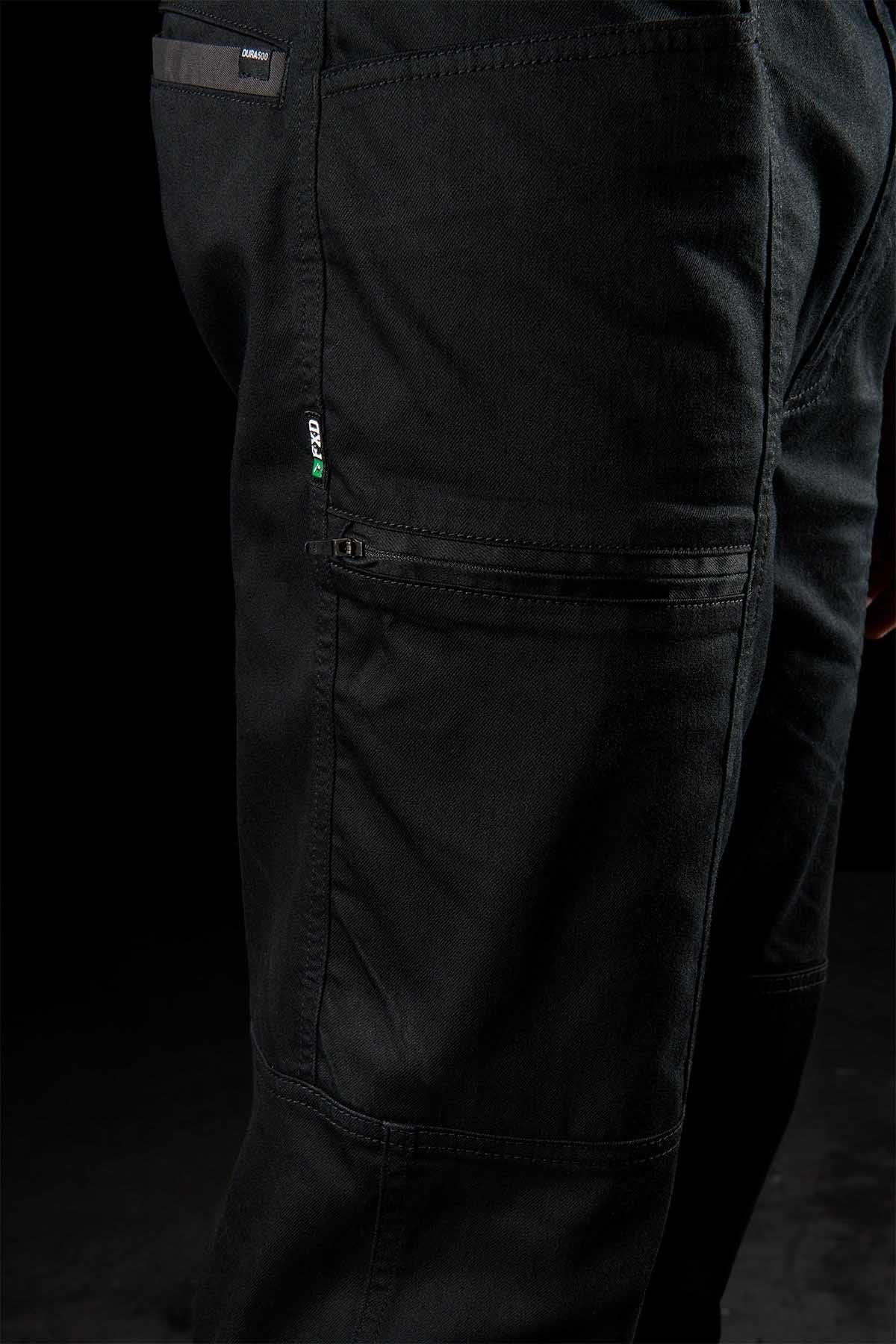 Close-up of FXD WP-6 black work pants highlighting a small green and gray logo tag on the side pocket. The fabric looks durable with reinforced stitching, featuring a stretch waistband and multiple pockets. These pants provide UPF50+ protection against UV rays. Dark background setting.