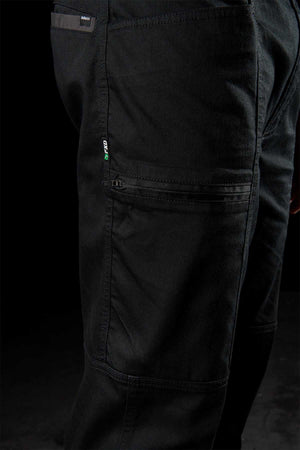 Close-up of FXD WP-6 black work pants highlighting a small green and gray logo tag on the side pocket. The fabric looks durable with reinforced stitching, featuring a stretch waistband and multiple pockets. These pants provide UPF50+ protection against UV rays. Dark background setting.