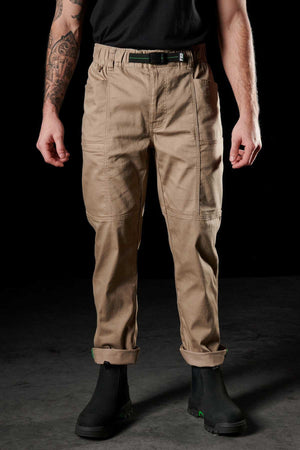 A person stands against a dark background wearing FXD WP-6 light brown work pants with stitched knee patches and stretch waistband, accessorized with a black belt, black boots, and displaying a tattooed left arm.