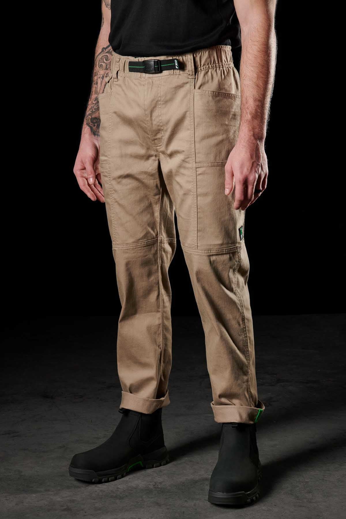 A person wears FXD WP-6 tan work pants with reinforced knees and a stretch waistband, along with a black belt, black t-shirt, and black work boots against a dark background. Only the torso and legs are visible.