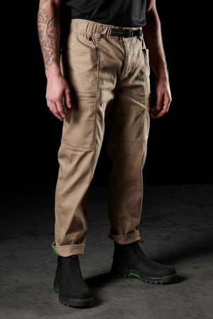 A person is seen from the waist down wearing FXD WP-6 tan work pants with a stretch waistband, rolled cuffs, a black belt, and black work boots. Visible tattoos are on one arm. The individual stands on a dark surface.