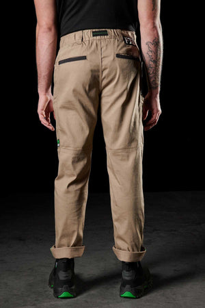 A person with a tattooed left arm wears FXD WP-6 beige work pants with stretch waistband and black pockets, paired with a black shirt and black shoes featuring green accents. The background is dark.