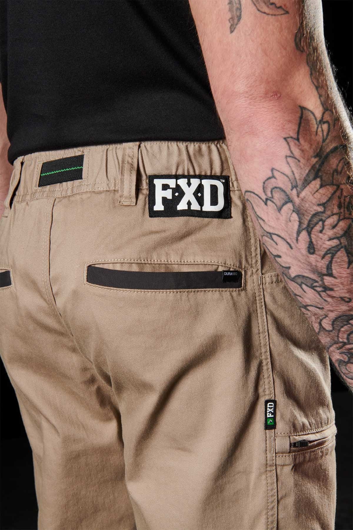 A person wears the FXD WP-6 work pant with a stretch waistband, black detailing, and the "FXD" patch above the back pocket. Their tattooed arm rests at their side against a dark background.