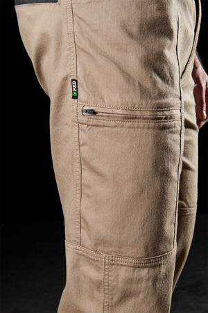 Close-up side view of the FXD WP-6 work pant with tan fabric, visible seams, stretch waistband, zippered pocket, and a small green and black CAT tag against a black background.
