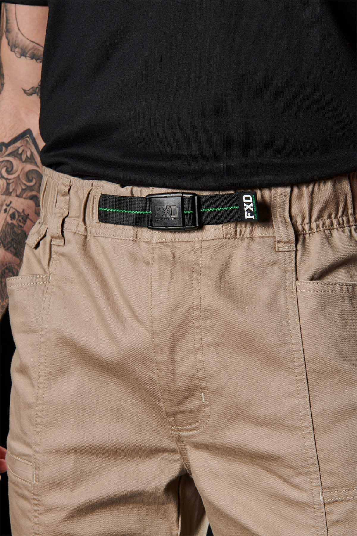 A person wears tan FXD WP-6 work pants with a black FXD belt and black shirt. The stretch waistband offers comfort, and part of a tattooed arm is visible on the left side of the image.
