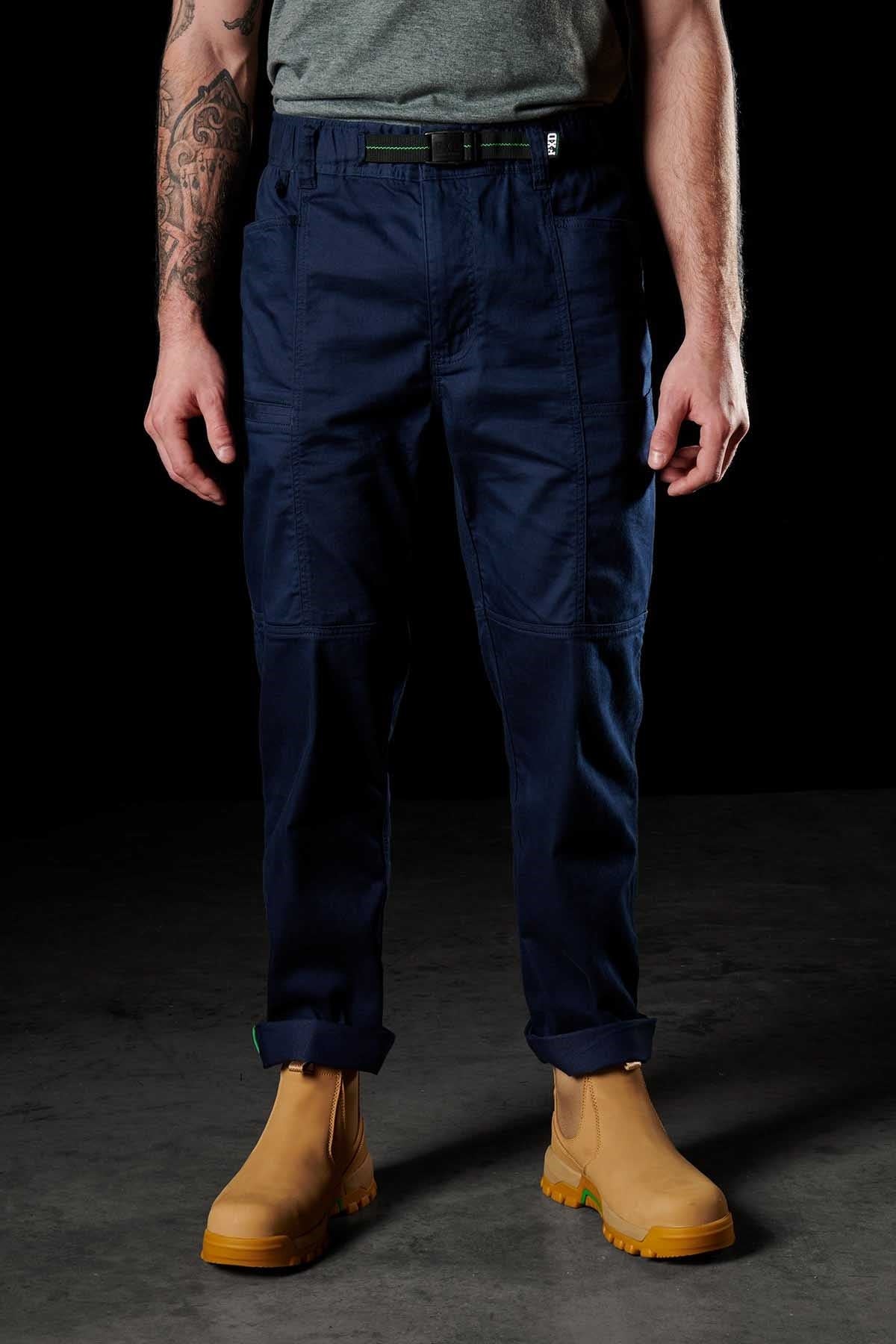 A person stands against a dark backdrop, wearing an FXD WP-6 gray shirt, durable work pants made from stretch nylon twill, and tan boots. Their tattooed arms hang relaxed by their sides.