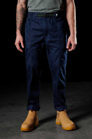 A person stands against a dark backdrop, wearing an FXD WP-6 gray shirt, durable work pants made from stretch nylon twill, and tan boots. Their tattooed arms hang relaxed by their sides.
