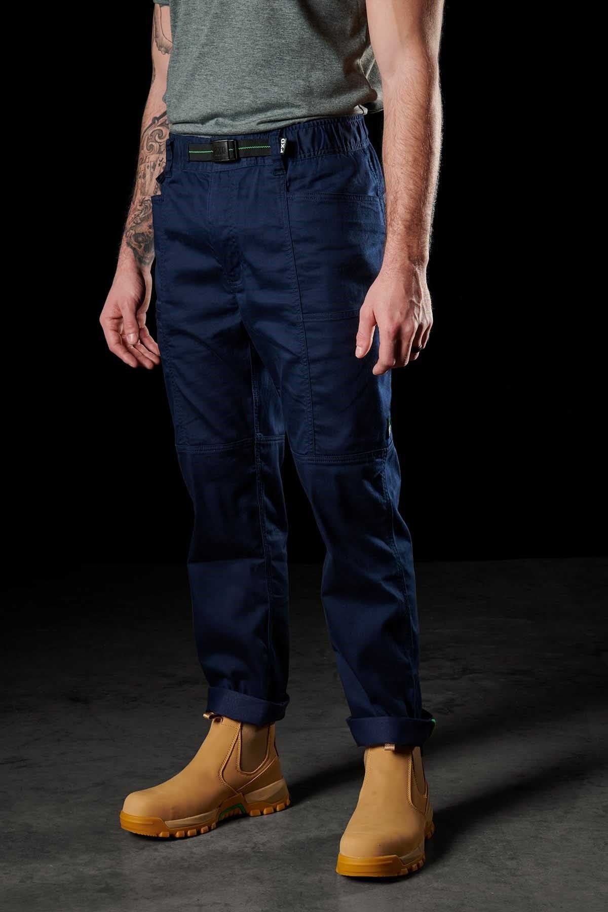Wearing FXD WP-6 blue pants made from stretch nylon twill, a gray T-shirt, and tan boots, the person stands against a dark background. Their forearm tattoo and hands in pockets enhance their casual, rugged style with these durable work pants.