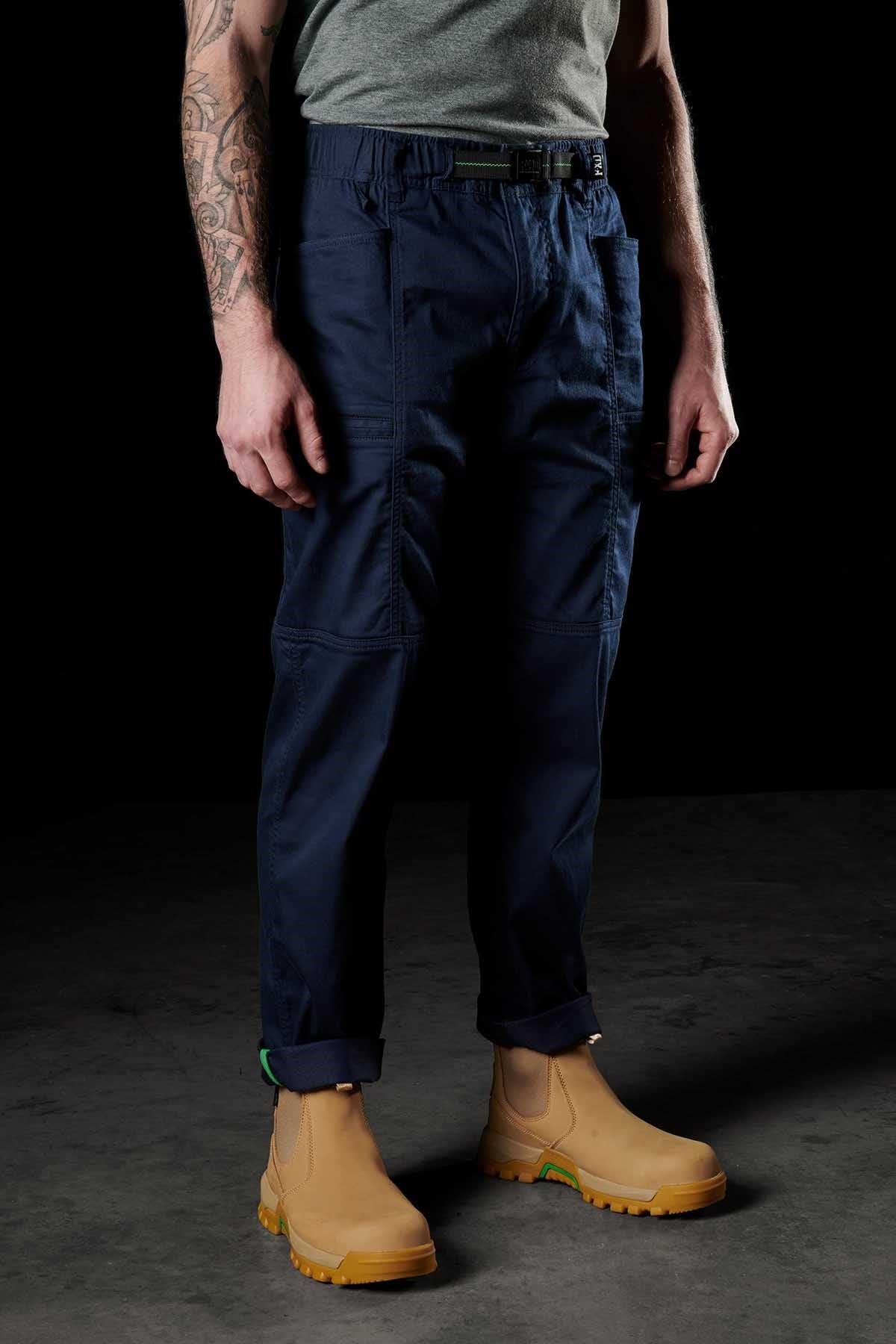 A person stands against a dark background wearing a gray t-shirt, blue FXD WP-6 cargo pants made from stretch nylon twill fabric, and tan work boots, showcasing a tattoo on their left arm.