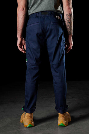 A person stands facing away, wearing a gray shirt and FXD WP-6 work pants in stretch nylon twill fabric. Tan boots with green soles complete the look, revealing tattoos on their left arm. The background is dark, and they are standing on concrete.