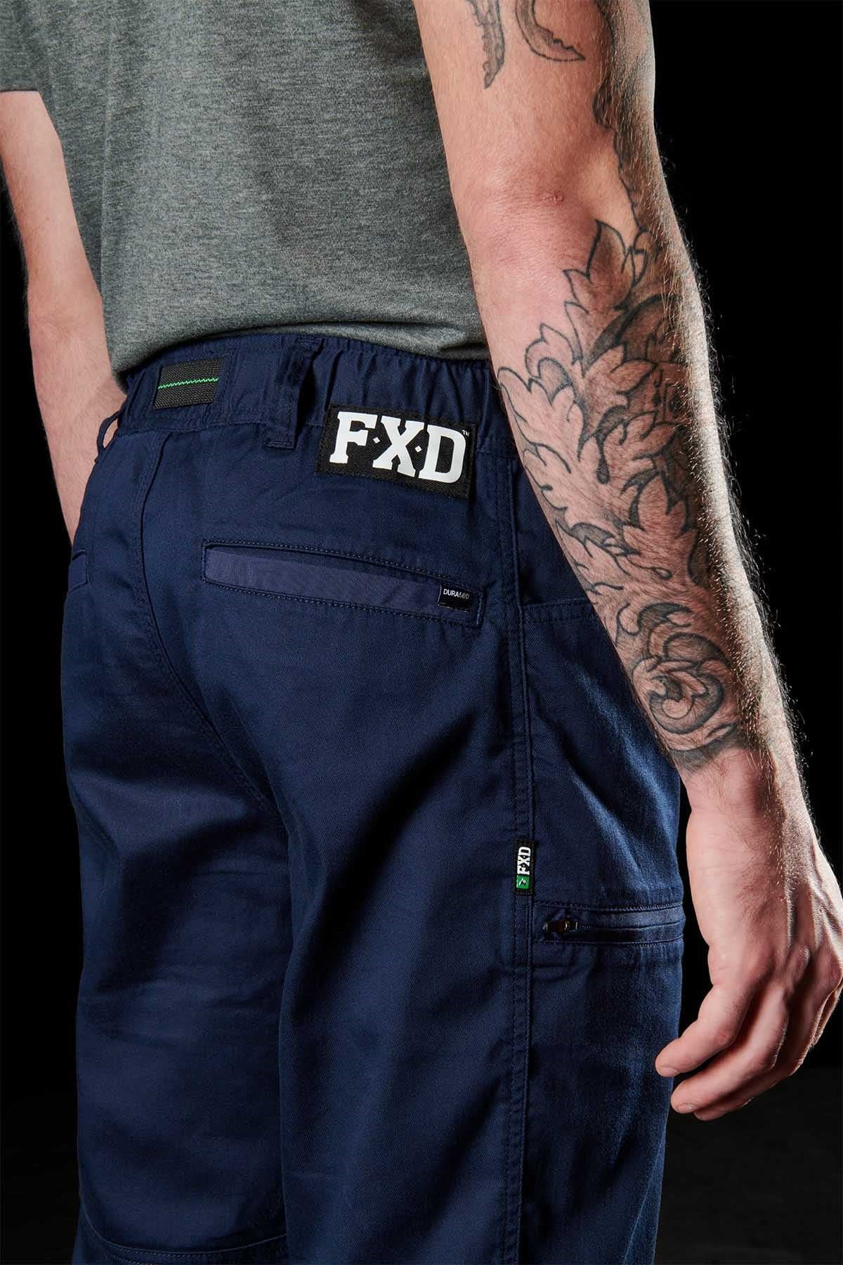 A person wears a grey t-shirt and FXD WP-6 work pants in navy blue, made from tough stretch nylon twill, with the logo on the waistband. A detailed floral tattoo adorns their right arm, and their left hand is in a pocket against a black backdrop.