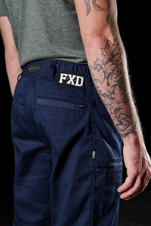 A person wears a grey t-shirt and FXD WP-6 work pants in navy blue, made from tough stretch nylon twill, with the logo on the waistband. A detailed floral tattoo adorns their right arm, and their left hand is in a pocket against a black backdrop.