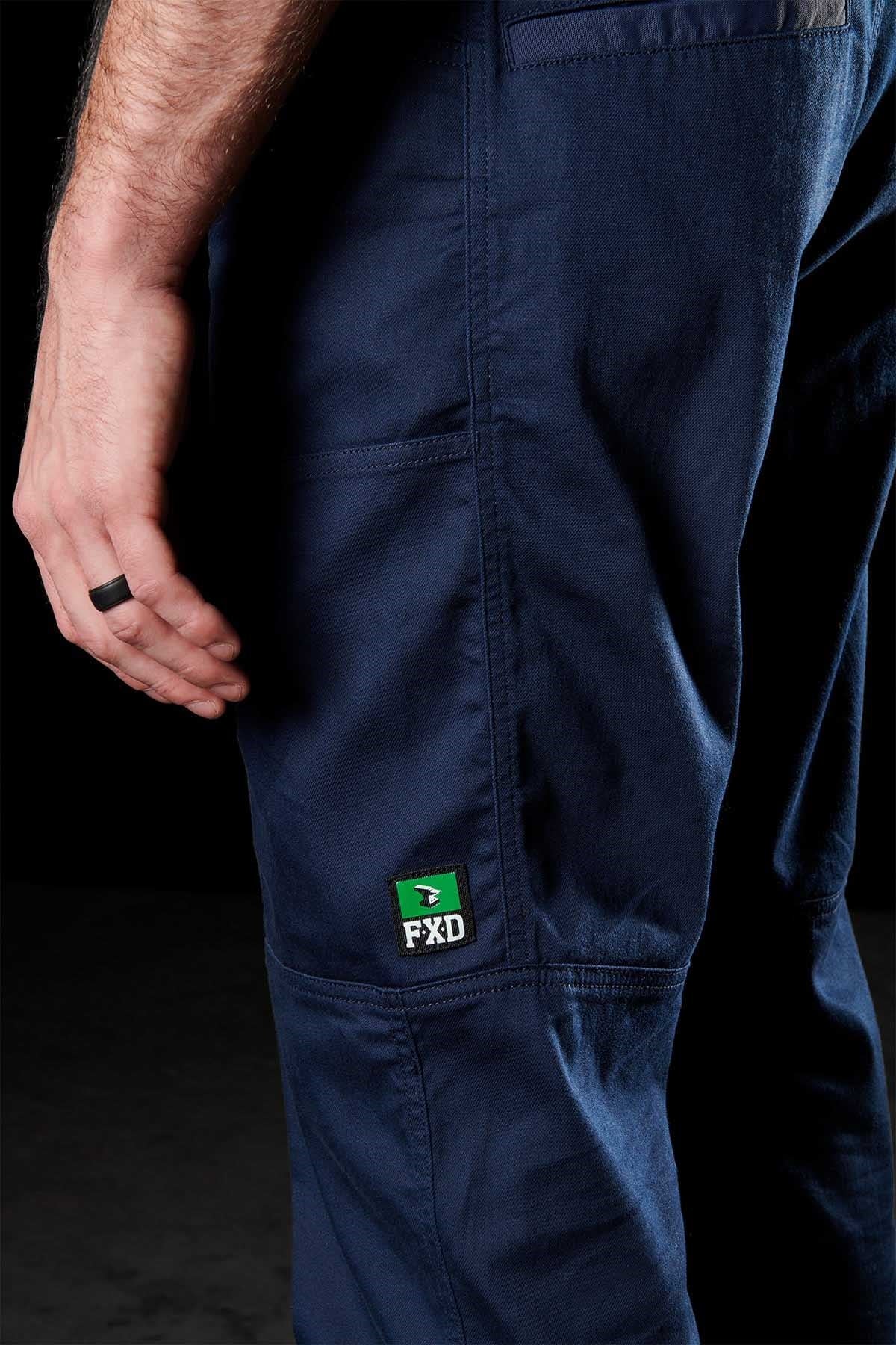 A person wears FXD WP-6 work pants made from stretch nylon twill, with the "FXD" label on the pocket. Their left hand, showing a black ring, is visible against a black background.
