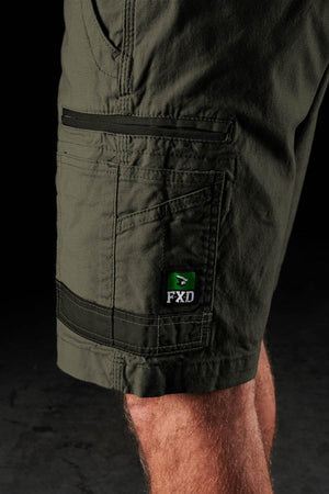 A close-up of a person wearing FXD WS-3 Short—durable stretch cotton canvas work shorts with contrast stitching, a side pocket, and an "FXD" logo patch above the knee. The background is dark and out of focus.