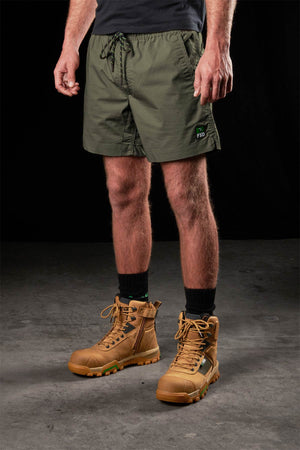 A person wearing FXD WS-4 olive green stretch ripstop work shorts, a black T-shirt, black socks, and tan lace-up boots stands on a concrete floor against a black background.
