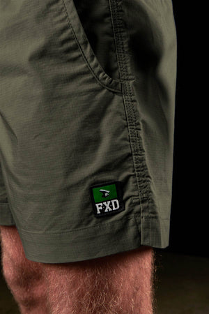 Close-up of a person wearing olive green FXD WS-4 Short made from stretch ripstop fabric, featuring a black and green FXD logo patch near the hem. Their hand is in the pocket, with the short standing out against a dark background.