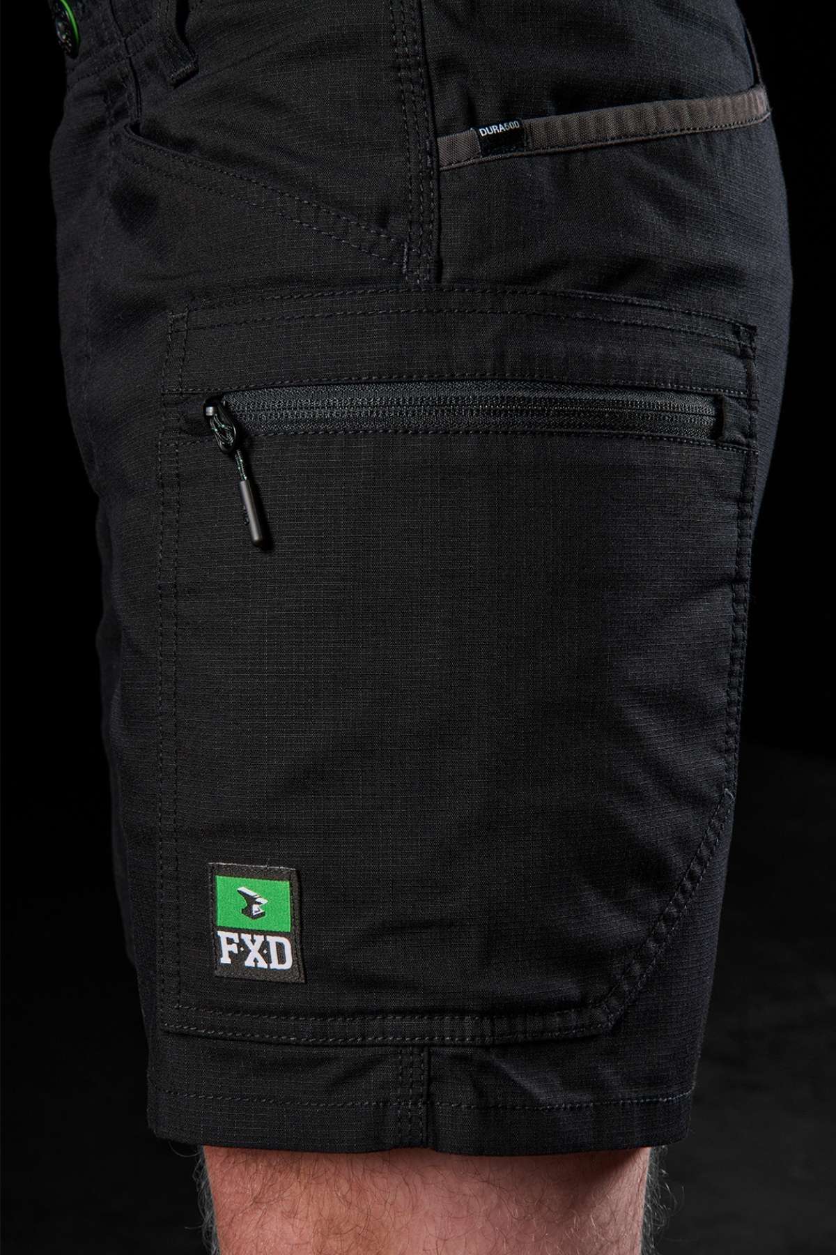 A close-up shot shows a person wearing the FXD WS-6 Work Short, featuring a zippered pocket and an FXD logo patch on the side. The dark background accentuates the stretch mobility and detailed design of these cargo shorts.