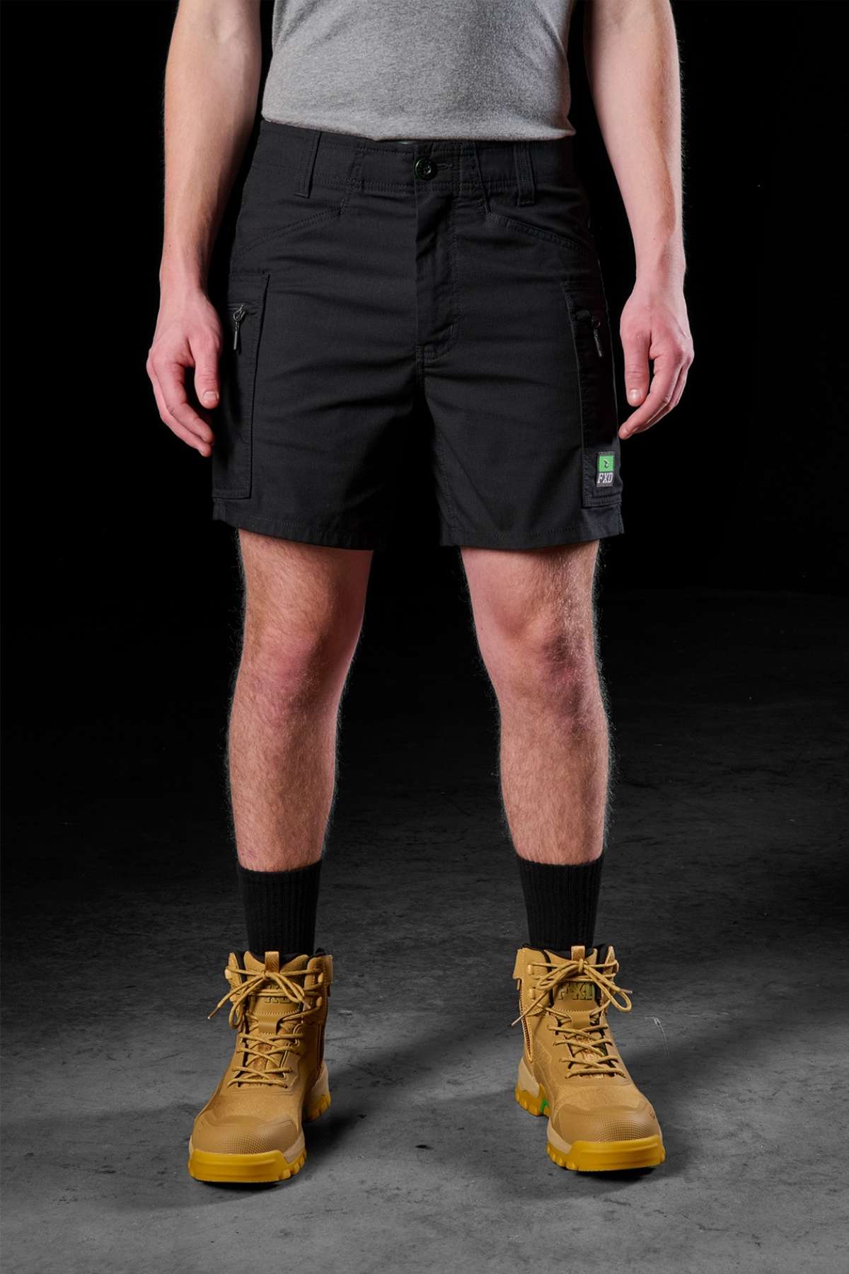 Against a dark backdrop, an individual displays the lower half of their attire, featuring the FXD WS-6 Work Short. These black cargo shorts are designed for stretch mobility and are paired with a gray shirt, black socks, and tan lace-up work boots.