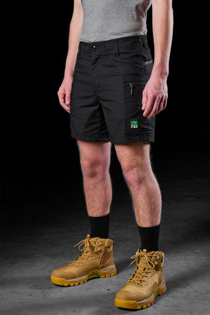 A person dressed in a gray shirt, black FXD WS-6 Work Short emblazoned with a patch, black socks, and tan work boots demonstrates the stretch mobility feature. The dark backdrop accentuates the practical style of the outfit.