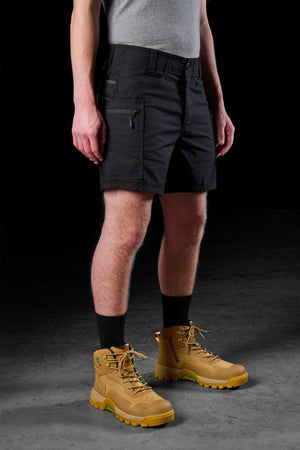 A person in a gray shirt, FXD WS-6 Work Short black cargo shorts, black socks, and tan hiking boots stands on a dark surface against a black background.