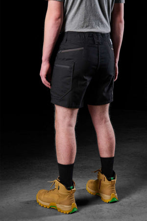 A person, facing away from the camera, is dressed in a gray t-shirt and the FXD WS-6 Work Short, known for its stretch mobility. They have black socks and yellow work boots on, standing against a dark background.