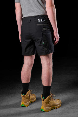 A person wearing a gray shirt, FXD WS-6 Work Shorts, and brown boots with green soles stands against a dark background. The stretch mobility of the shorts, featuring the "FXD" branding, accentuates comfort and style in this rear-view shot.