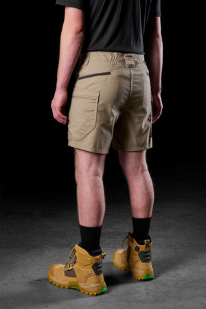 Wearing a black shirt, FXD WS-6 Work Shorts in khaki, black socks, and tan work boots with green soles, a person stands against a dark background. The view from behind showcases the outfit’s comfort and practicality.